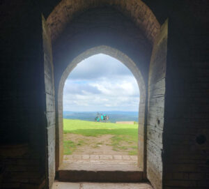 Vue sur la porte ouverte de l'église depuis l'intérieur. Un halo de lumière se dessine dans l'arche de la porte et au loin, on aperçoit un vélo équipé de sacoche de cylotourisme. 