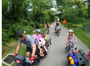 Un père de famille pédale sur un vélo long tail avec deux jeunes enfants à l'arrière pendant des vacances à vélo. Une petite fille pédale seule, sa maman la suit. 