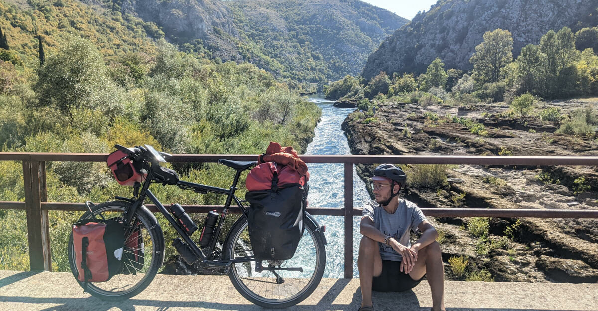 Pierre est auteur du blog En Roue libre, il est assis à côté de son vélo, sur un pont, avec une rivière derrière lui.