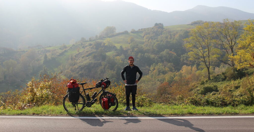 Pierre, l'auteur du blog En Roue Libre, se tient debout à côté de son vélo équipé de sacoches, devant un paysage de forêt en automne. 