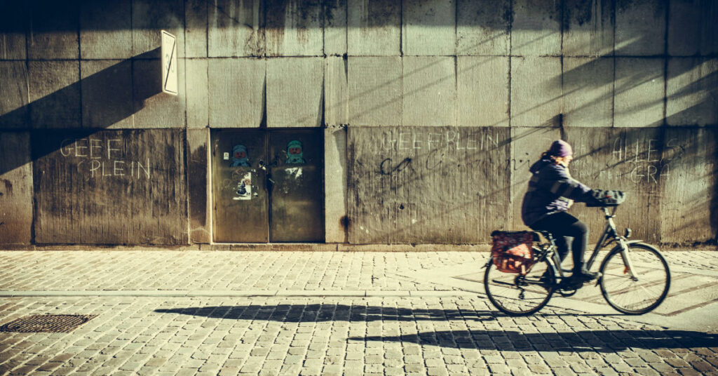 Un cycliste est à droite de l'image et roule sur un vieux vélo avec des sacoches devant une vieille façade d'immeuble taguée. 