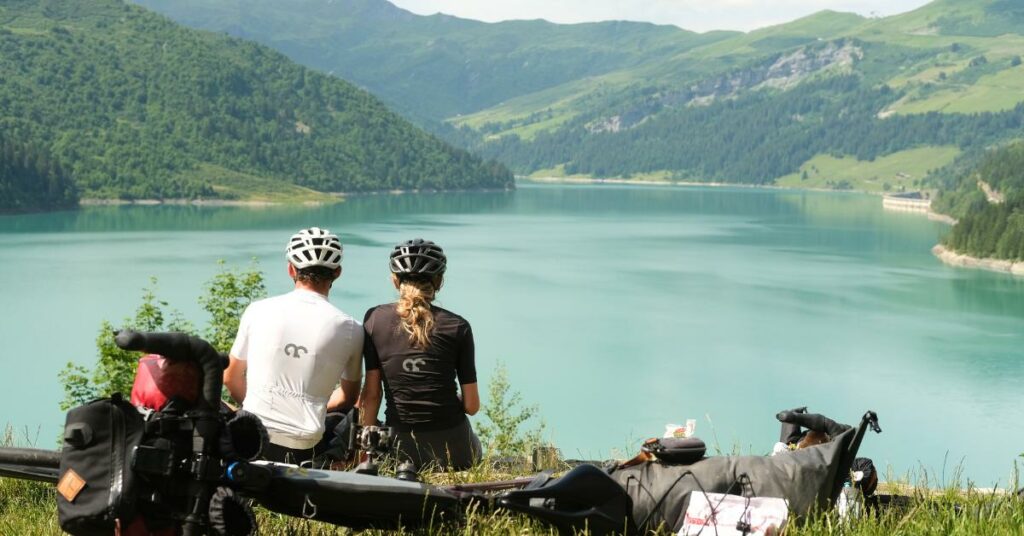 Amaury et Denisa sont tous les deux assis dans l'herbe, face au lac, avec leur vélo à côté. 