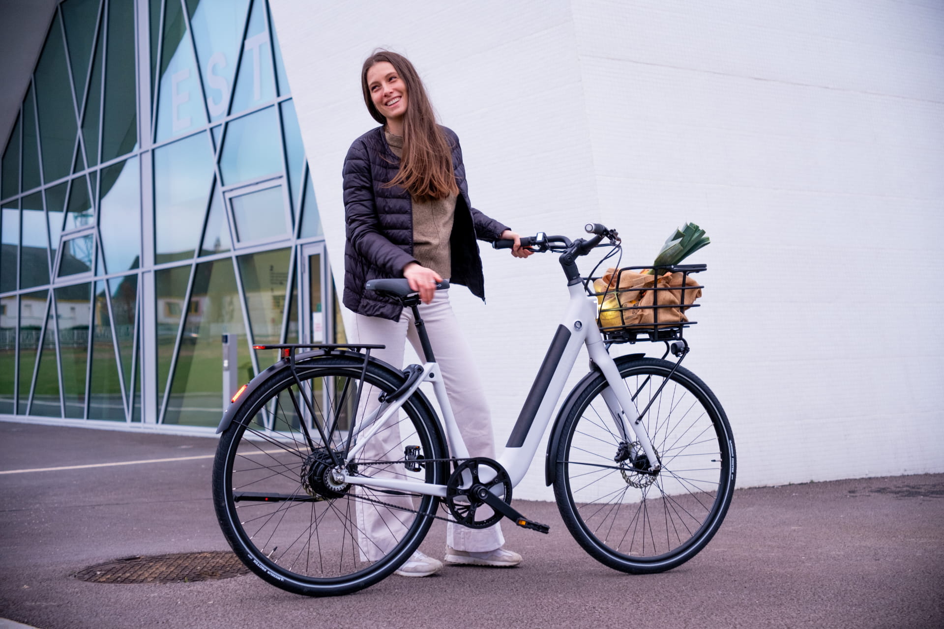 une jeune femme tient un vélo électrique Ellipse Bike avec un panier avant contenant des légumes. Elle porte une doudoune noire et le vélo est blanc